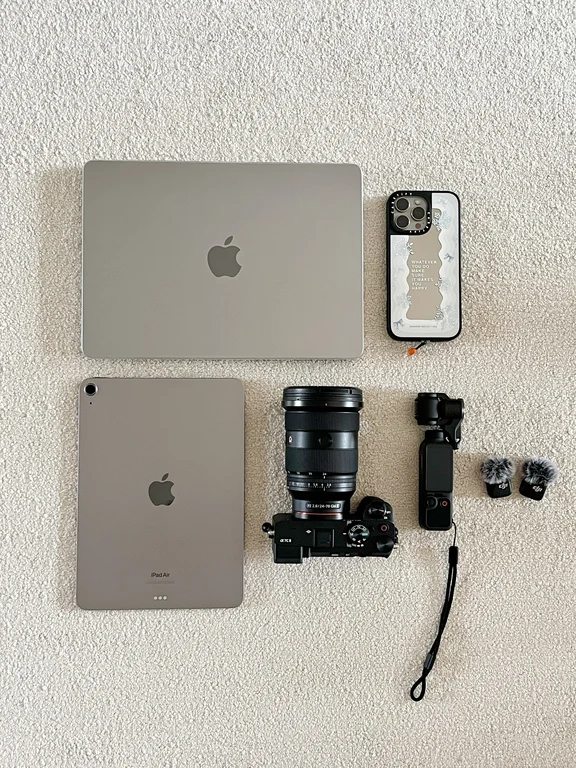 Flat lay of Apple tech gear including a MacBook, iPad Air, iPhone in a case, a camera with zoom lens, and audio equipment