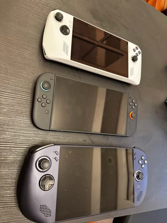 Three handheld gaming devices lined up on a dark desk including a white ROG Ally and a dark console