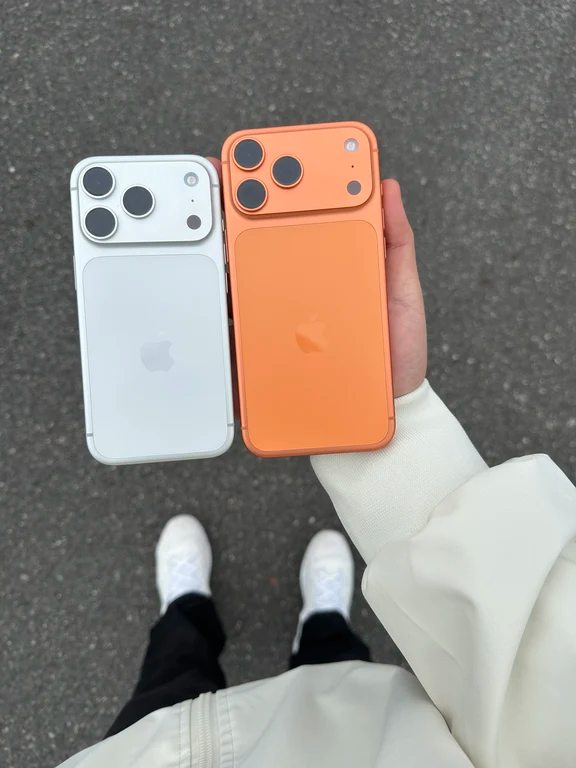 Hand holding two smartphones side-by-side, one white and one bright cosmic orange, displaying their redesigned rear camera island visor.