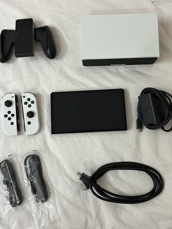 Nintendo Switch OLED white console, dock, Joy-Con controllers, grip, and cables laid out on a sheet