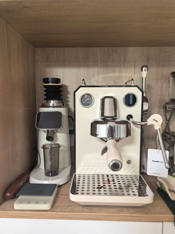 White Gemilai espresso machine with portafilter and pressure gauge next to an electric grinder on a wooden kitchen counter