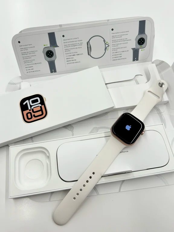 Unboxed Apple Watch with a starlight gold case and white sport band resting on packaging showing the Apple logo on screen