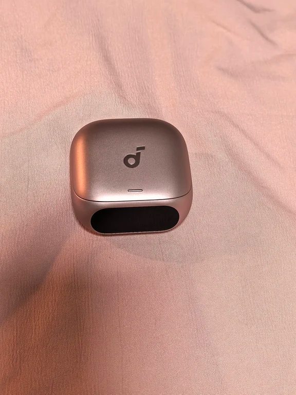 Metallic bronze Soundcore earbud case sitting closed on soft pink fabric