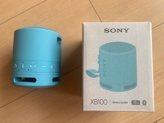 Light blue Sony SRS-XB100 wireless portable speaker next to its cardboard retail box showing 16h battery mark.