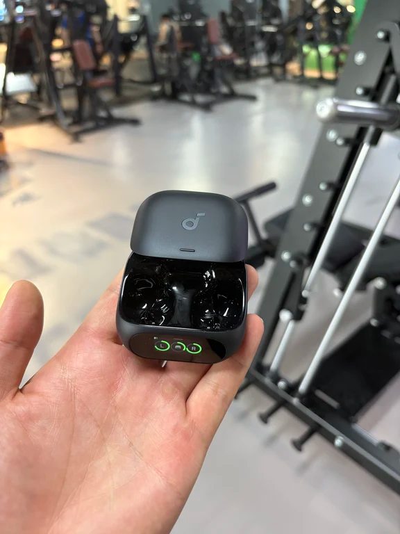 Hand holding open black Soundcore charging case displaying battery levels on a small front screen in a gym