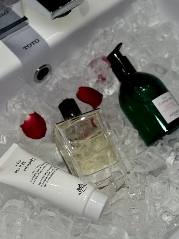 Hermès Les Mains Hermès hand cream tube, perfume bottle, and green Eau d'Orange Verte bottle resting over ice cubes with rose petals.