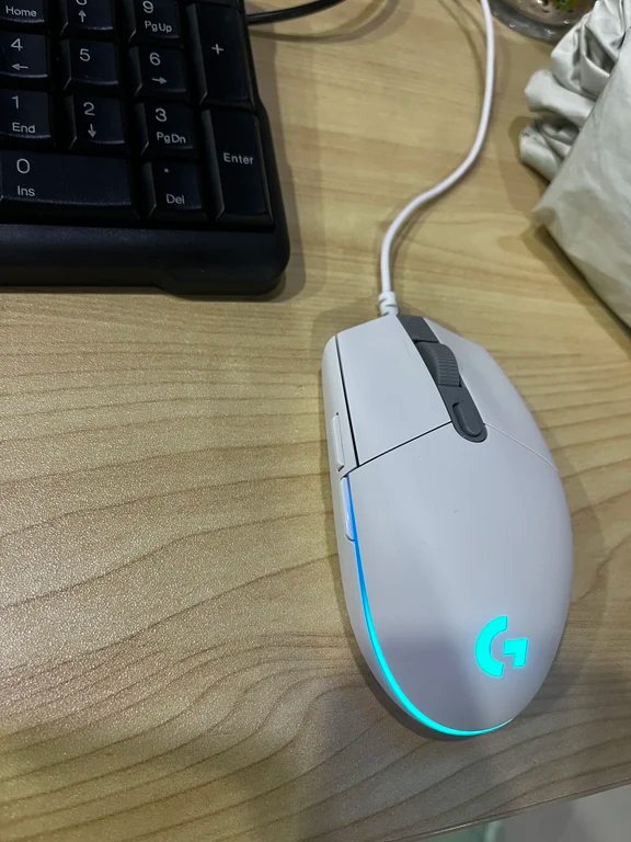 White Logitech G102 mouse powered on with cyan RGB illumination along the base