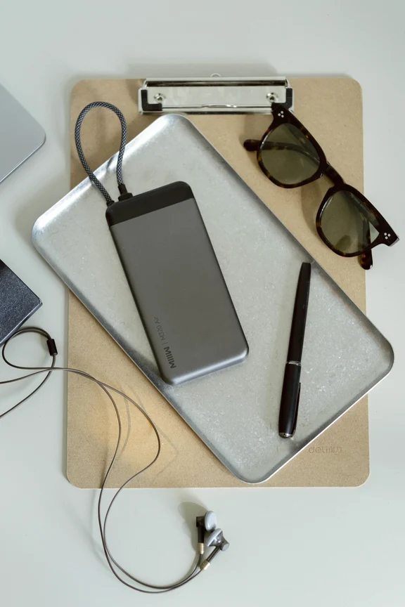 A slim dark gray MIIIW power bank with an integrated strap placed on a silver metal tray next to a black pen and sunglasses.
