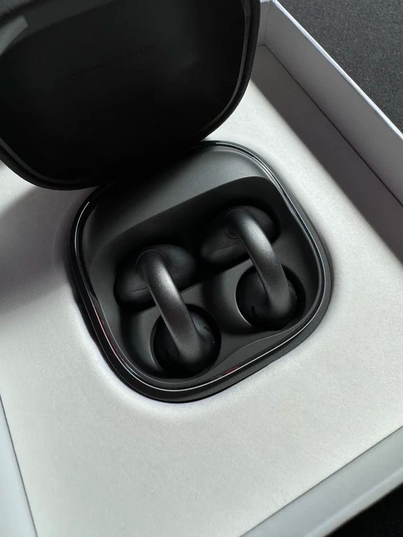 Matte black ear-clip wireless earphones nested in an open charging case inside original packaging.