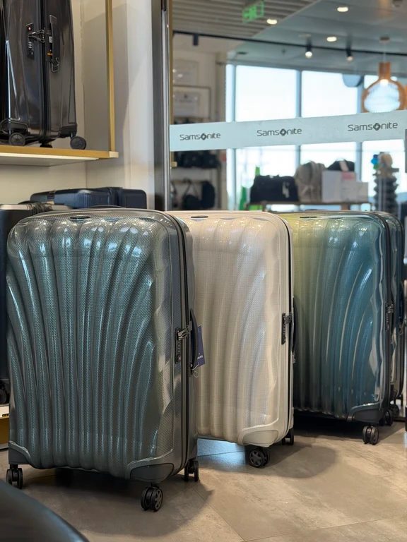 Three Samsonite hardside suitcases in metallic green, pearl white, and teal standing in a luggage store