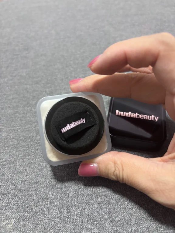 Hand holding a compact travel-size Huda Beauty loose powder jar with black sponge applicator