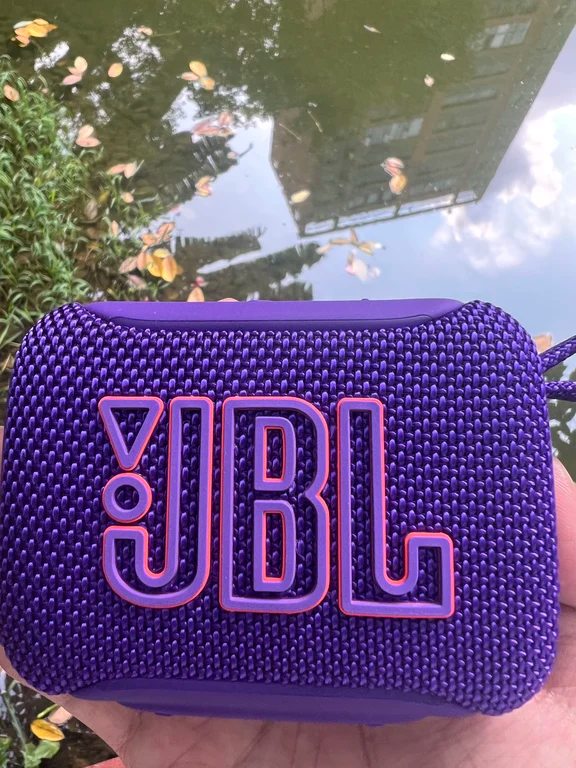 Purple JBL compact outdoor speaker held in hand showing rugged textured woven grille and carry loop.