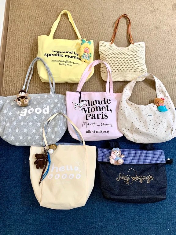 Flat lay of six colorful canvas tote bags in yellow, pink, grey, and cream on carpet