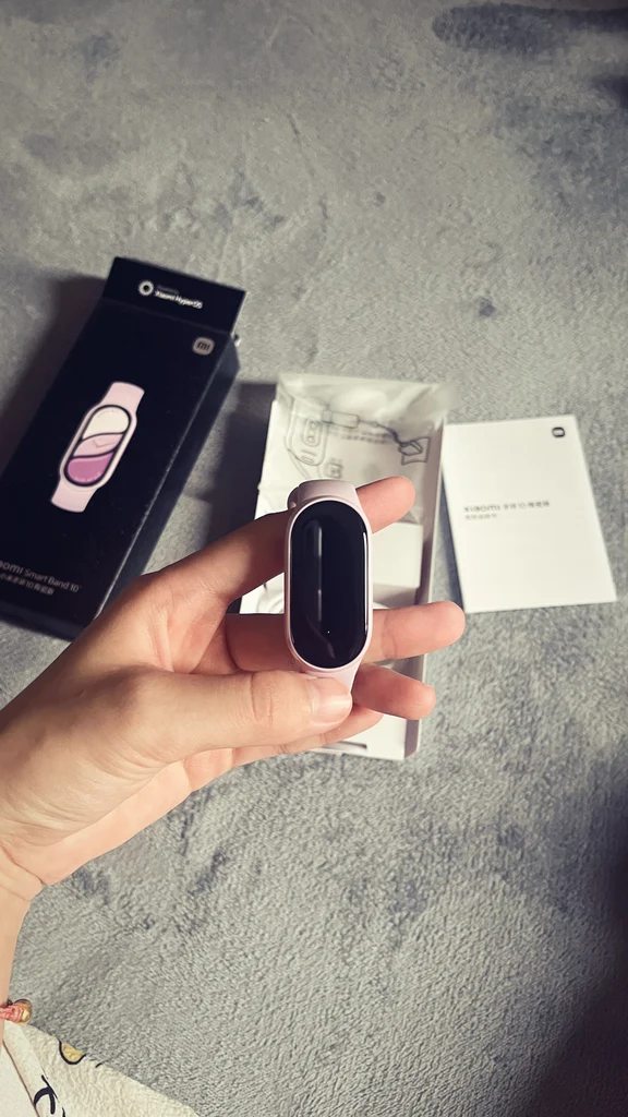 A hand holding a compact oval fitness tracker with a light pink band in front of its retail box