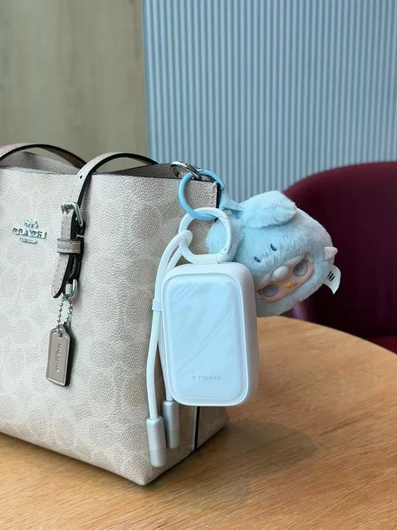 A compact white Torras power bank with hanging cables clipped onto the handle of a Coach handbag next to a blue plush toy.
