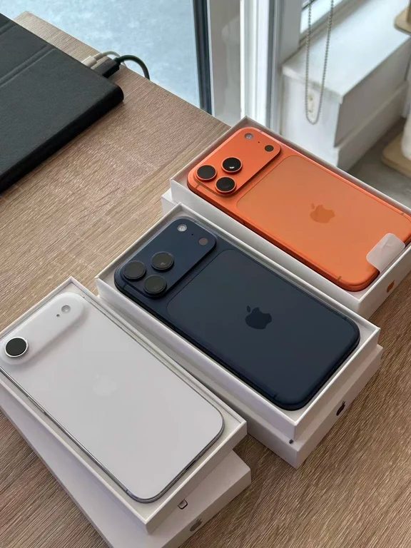 Three smartphones placed inside their white retail boxes side by side, displaying white, dark blue, and orange finishes.