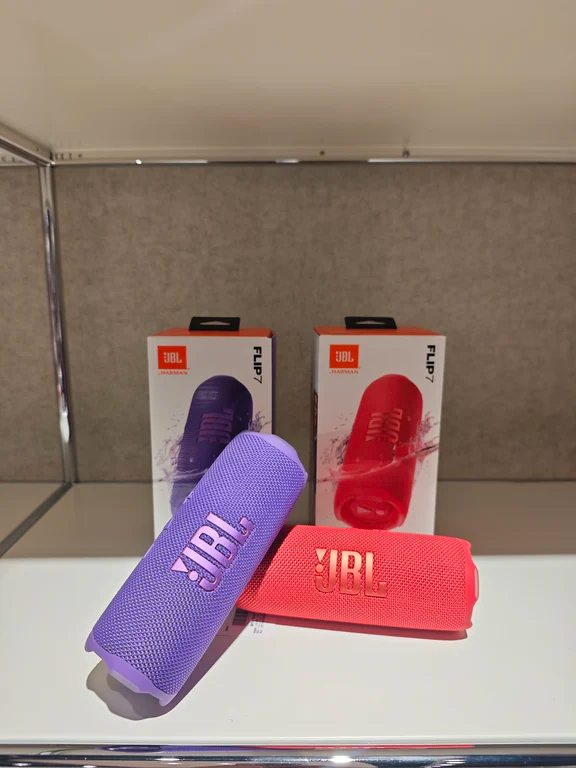 Two JBL Flip 7 portable Bluetooth speakers in purple and red positioned in front of their official product packaging.