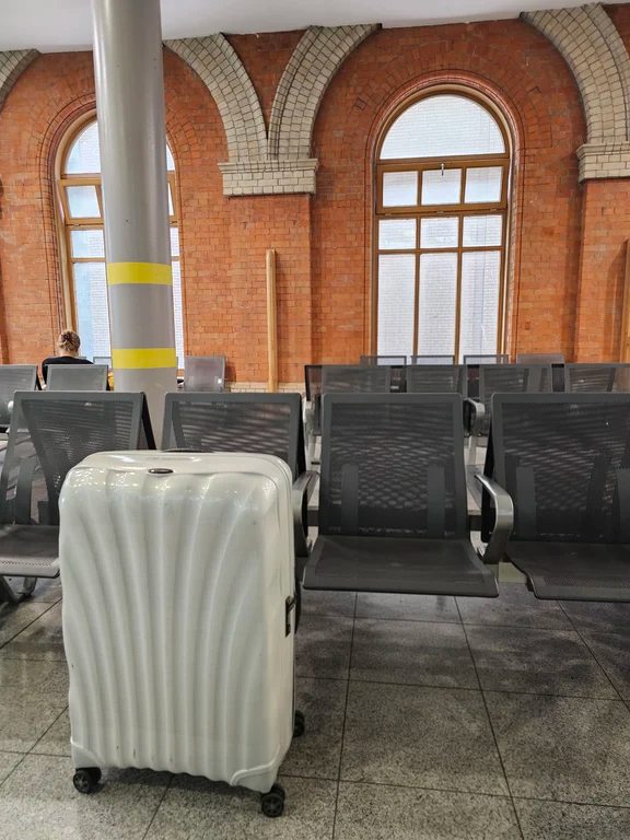 A white textured Samsonite carry-on suitcase placed beside waiting lounge chairs in a transit hall