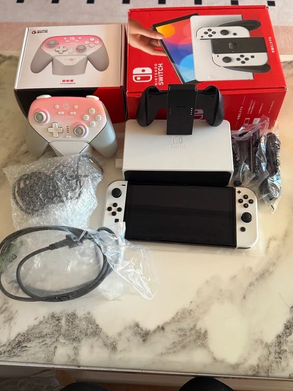 Nintendo Switch OLED tablet with white Joy-Cons, dock, red retail box, and a pink controller on a marble surface