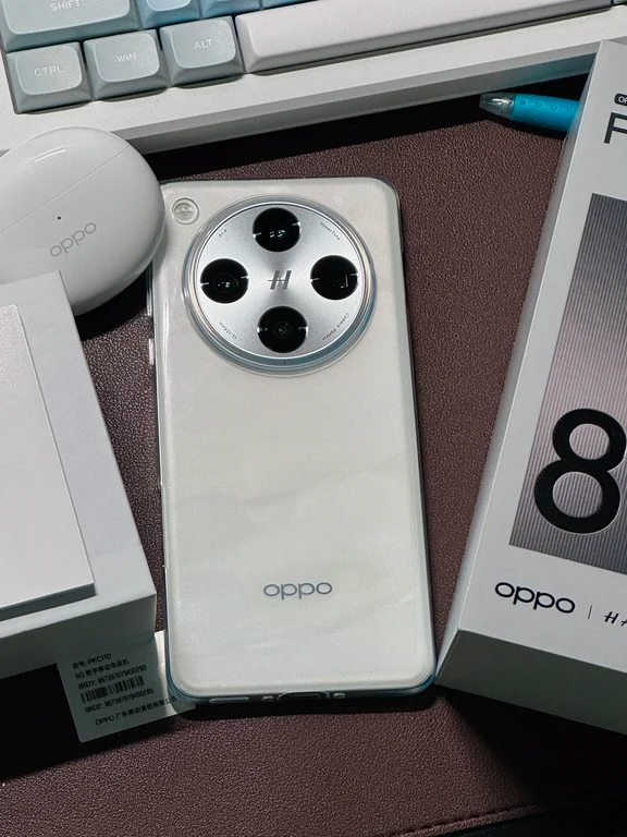 White smartphone featuring a large circular camera module with Hasselblad branding, shown next to an earphone case and product box on a desk