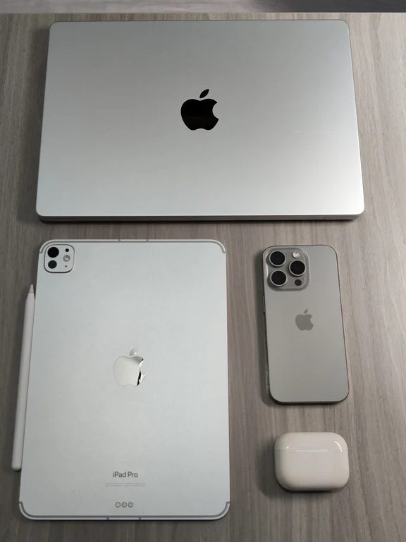Apple devices arranged on a desk including a MacBook, an iPad Pro with Apple Pencil, an iPhone, and AirPods.