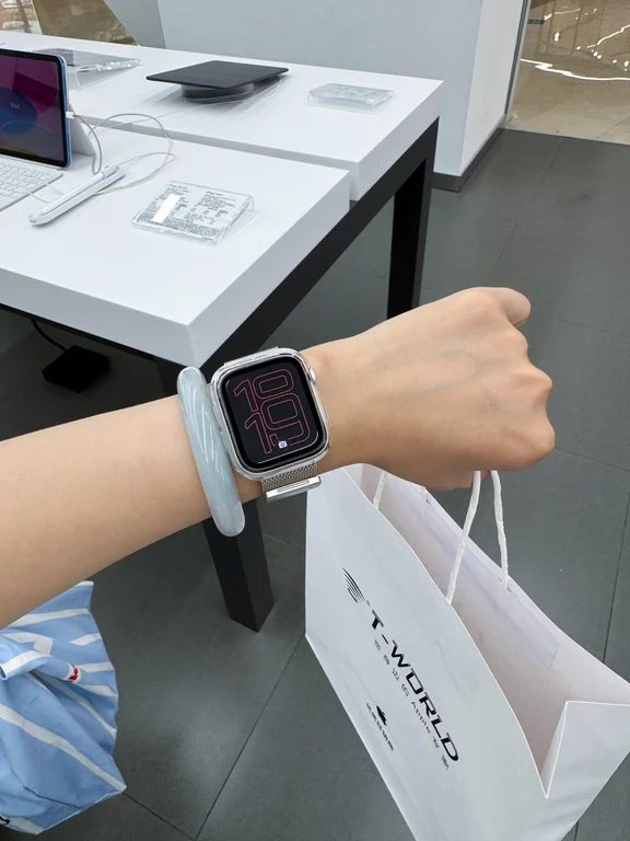 A person wearing a silver smartwatch with a digital clock face next to a shopping bag inside a retail store