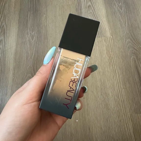 Hand holding a bottle of Huda Beauty liquid foundation displaying the shade through the clear container