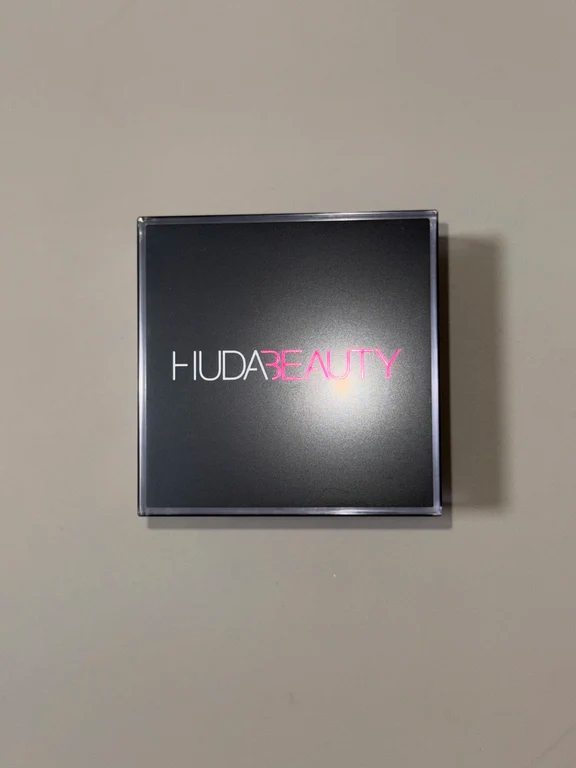 Square black makeup compact case with Huda Beauty logo printed in pink letters