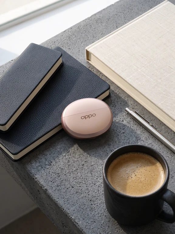Rose gold Oppo earbud charging case lying beside notebooks, a metal pen, and a coffee mug